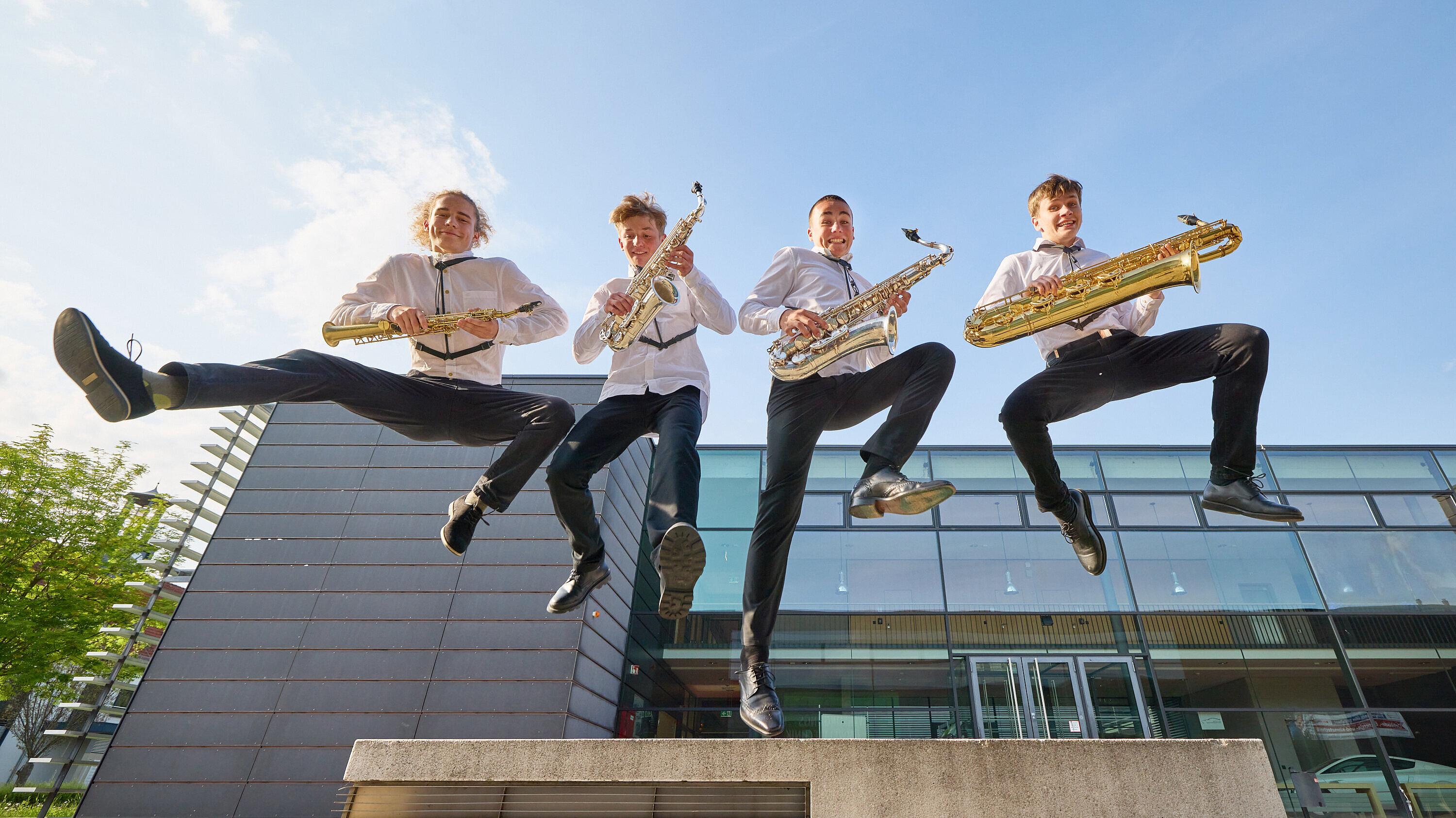 Vier Jugendliche mit je einem Saxophon in der Hand springen in die Luft.