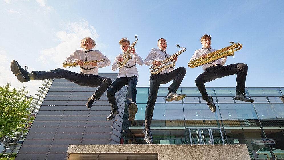 Vier Jugendliche mit je einem Saxophon in der Hand springen in die Luft. Im Hintergrund eine Glasfront und blauer Himmel.