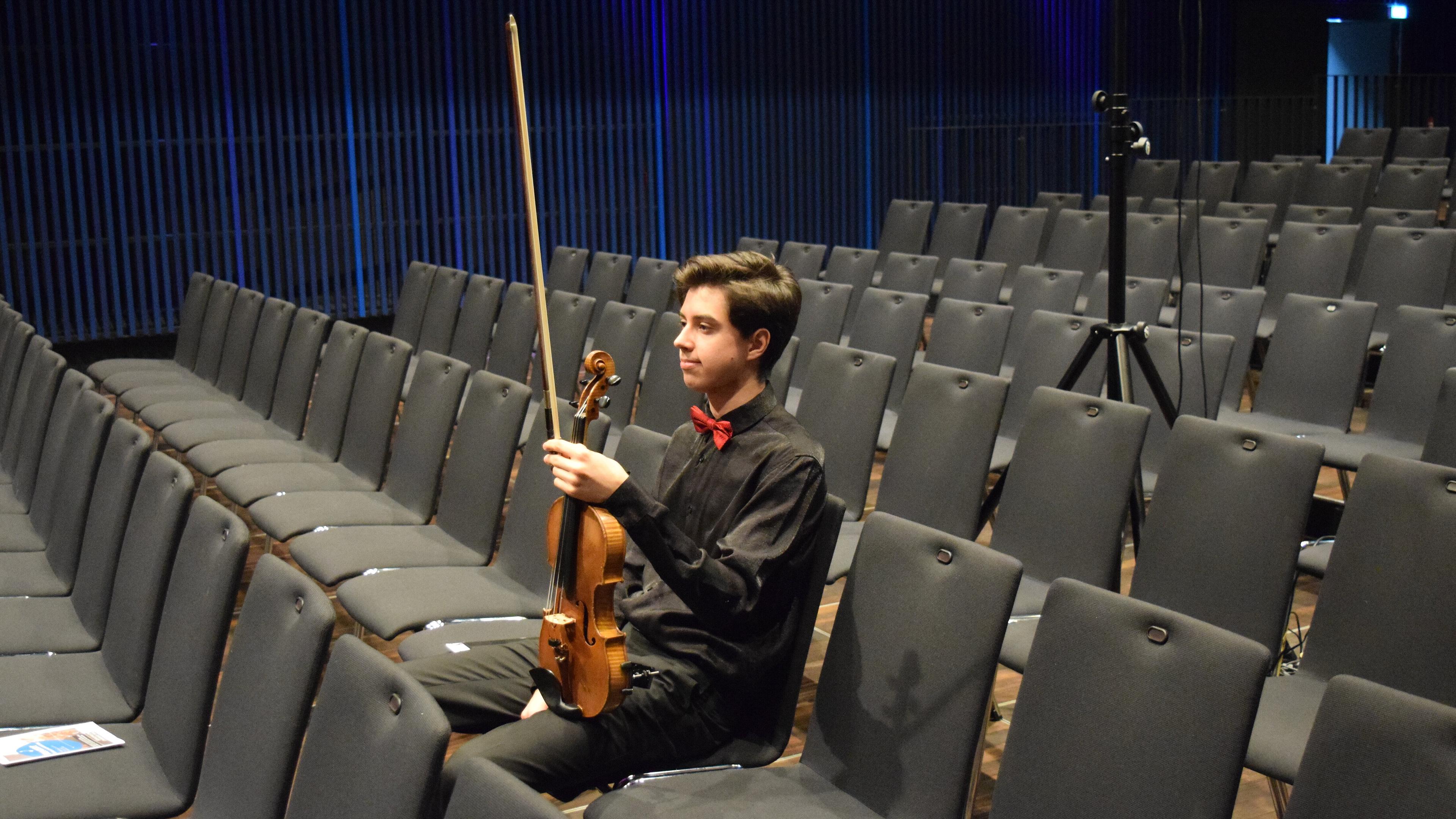 Jugendliche Person in Konzertkleidung hält eine Geige in der Hand, sitzt allein in leeren Stuhlreihen und wartet.