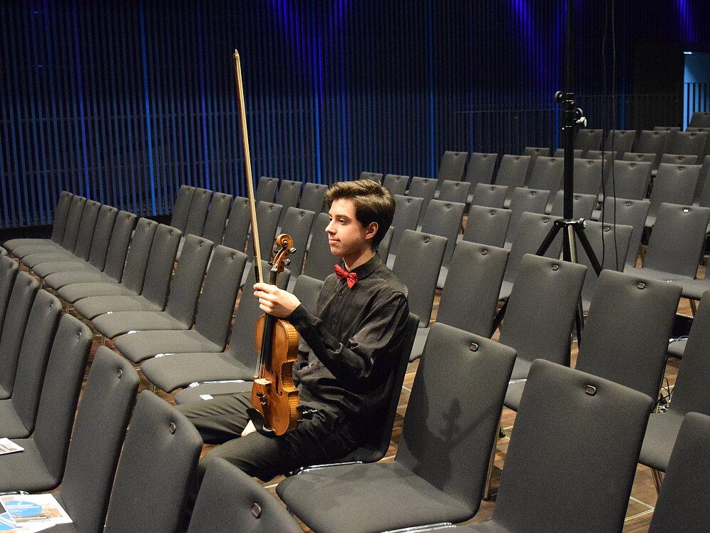 Jugendliche Person in Konzertkleidung hält eine Geige in der Hand, sitzt allein in leeren Stuhlreihen und wartet.