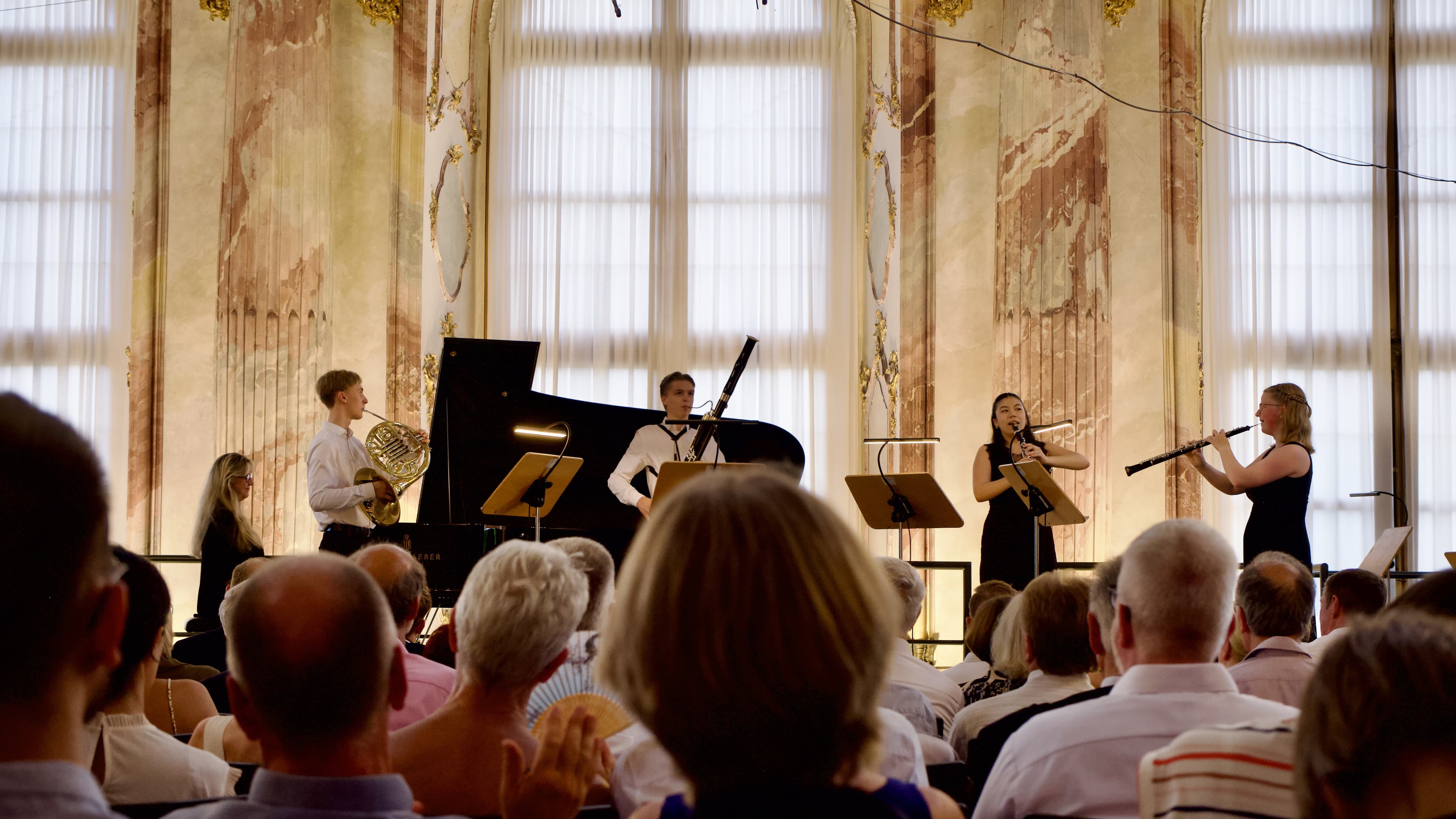 Klavierquintett während des Konzerts. Die Hinterköpfe des Publikums sind im Vordergrund, im Hintergrund die stuckverzierte Wand mit bodentiefen Rundbogenfenstern.