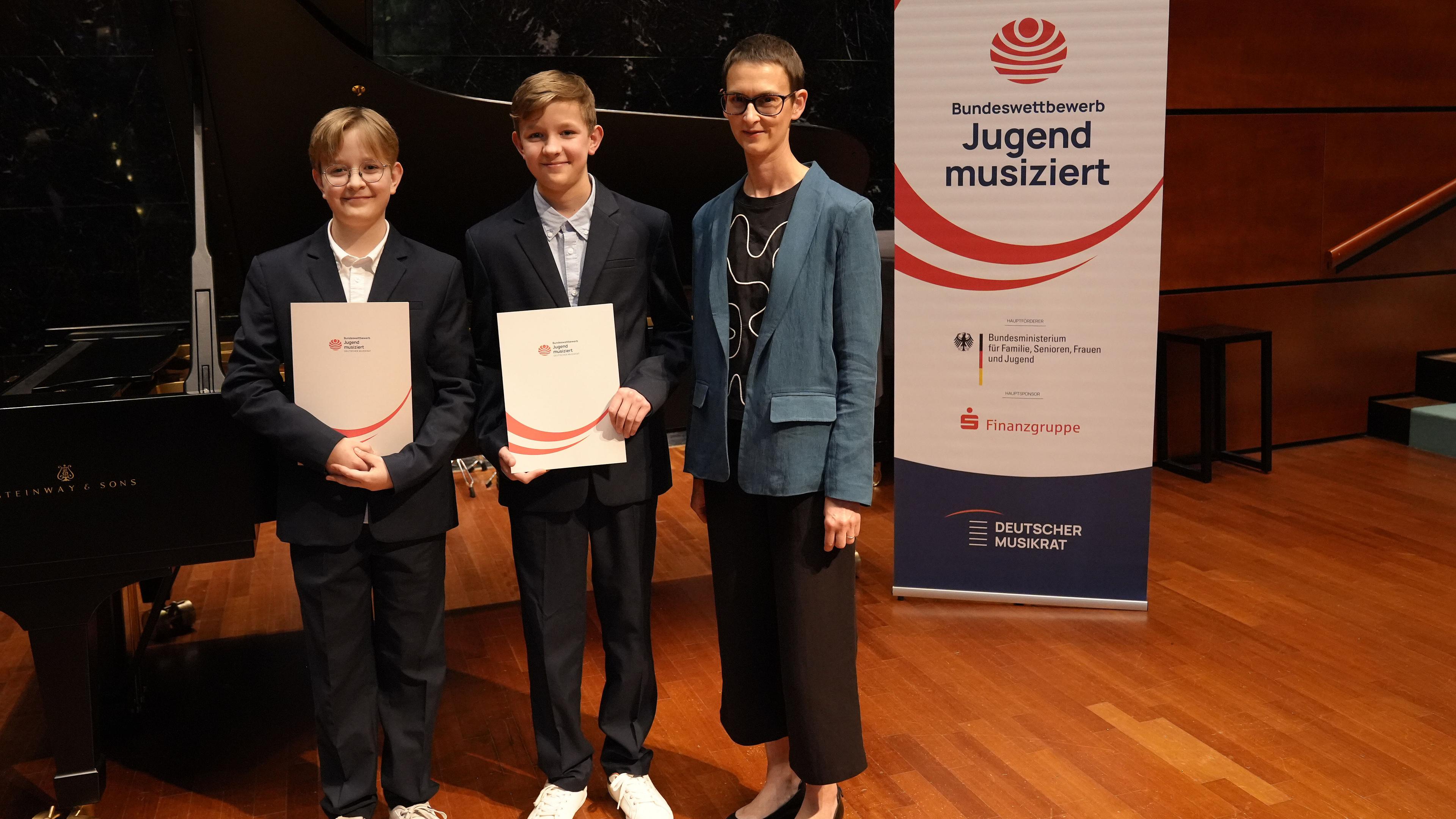 Die Arestovs stehen mit Ulrike Kniesner vor einem Flügel und zeigen lächelnd ihre Urkunden.